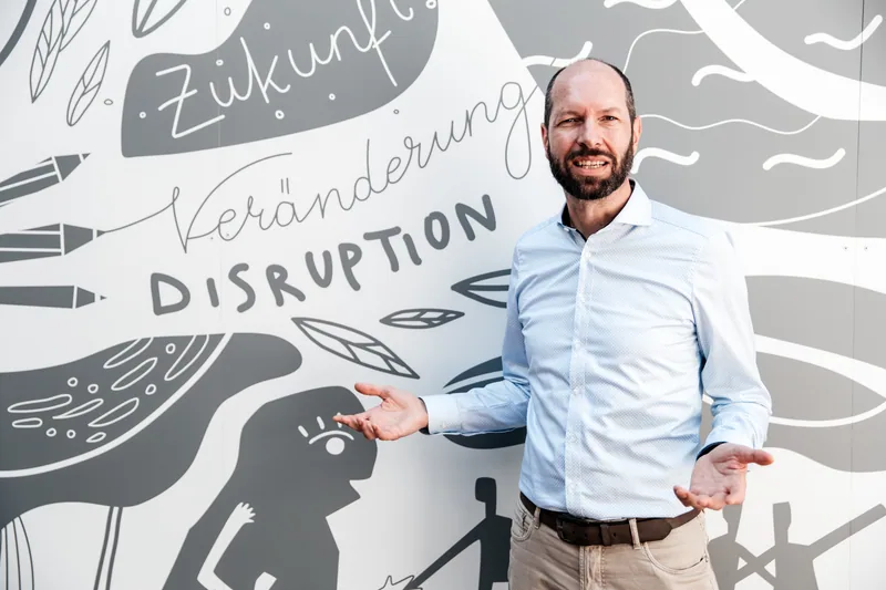 Georg Fischer steht vor einer Wandillustration mit den Worten Zukunft, Veränderung und Disruption und weist mit offener Hand darauf.