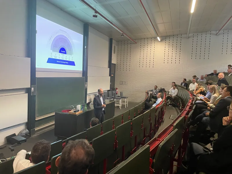 Ein Hörsaal mit ansteigenden Bankreihen, vorne eine Projektion, einzelne Zuhörende verteilt.