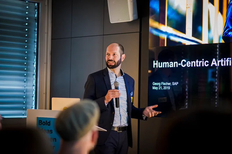 Georg Fischer spricht mit Mikrofon auf einer Bühne vor einer blauen Projektion zum Thema Human-Centered.
