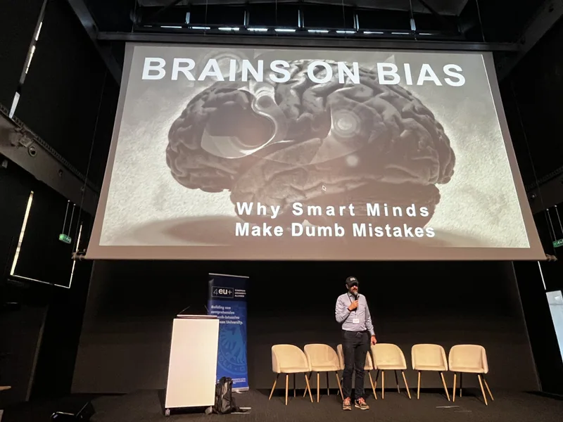 Eine Bühne mit der Projektion "Brains on Bias — Why Smart Minds Make Dumb Mistakes", davor der Redner.